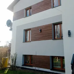 Nowoczesny dom z elewacją w odcieniach szarości i brązu, z oknami w drewnianych ramach, oświetleniem zewnętrznym i anteną satelitarną na balkonie.