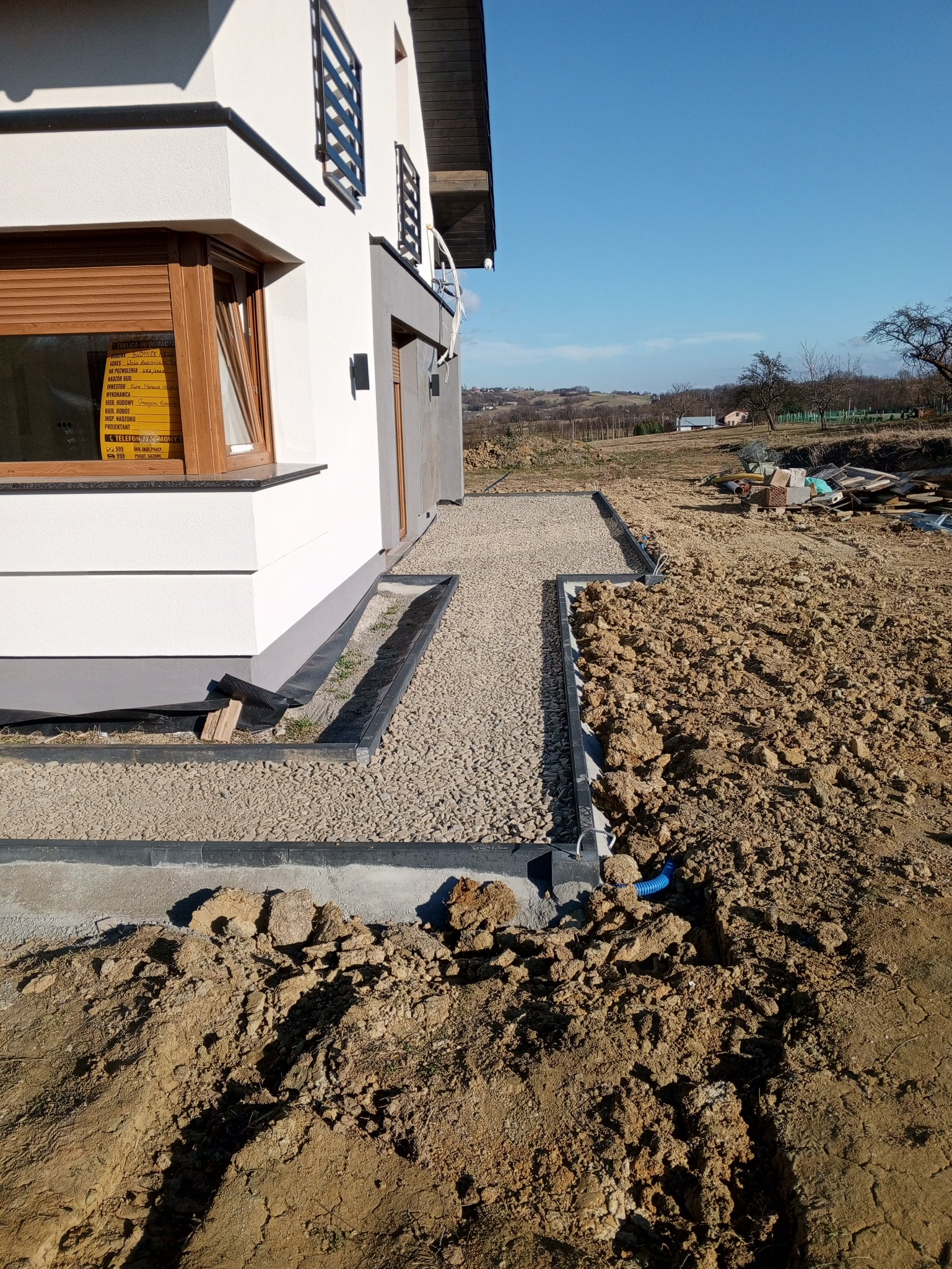 Ścieżka z jasnego żwiru obramowana czarnym obrzeżem, prowadząca wzdłuż ściany domu z widoczną tablicą informacyjną budowy, w tle krajobraz z zabudowaniami.