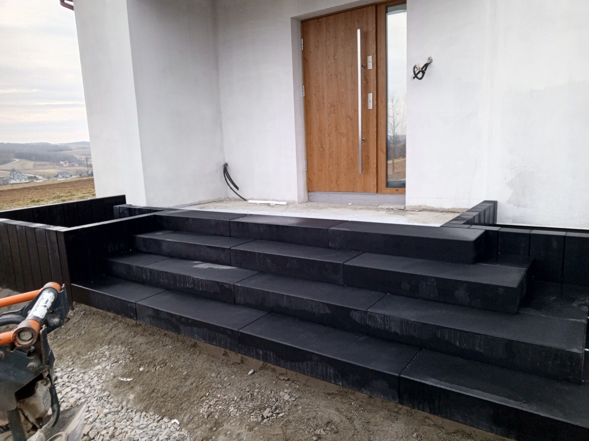 Nowoczesne, czarne schody wejściowe prowadzące do domu z drewnianymi drzwiami i białymi ścianami. W tle widoczny pagórkowaty krajobraz.