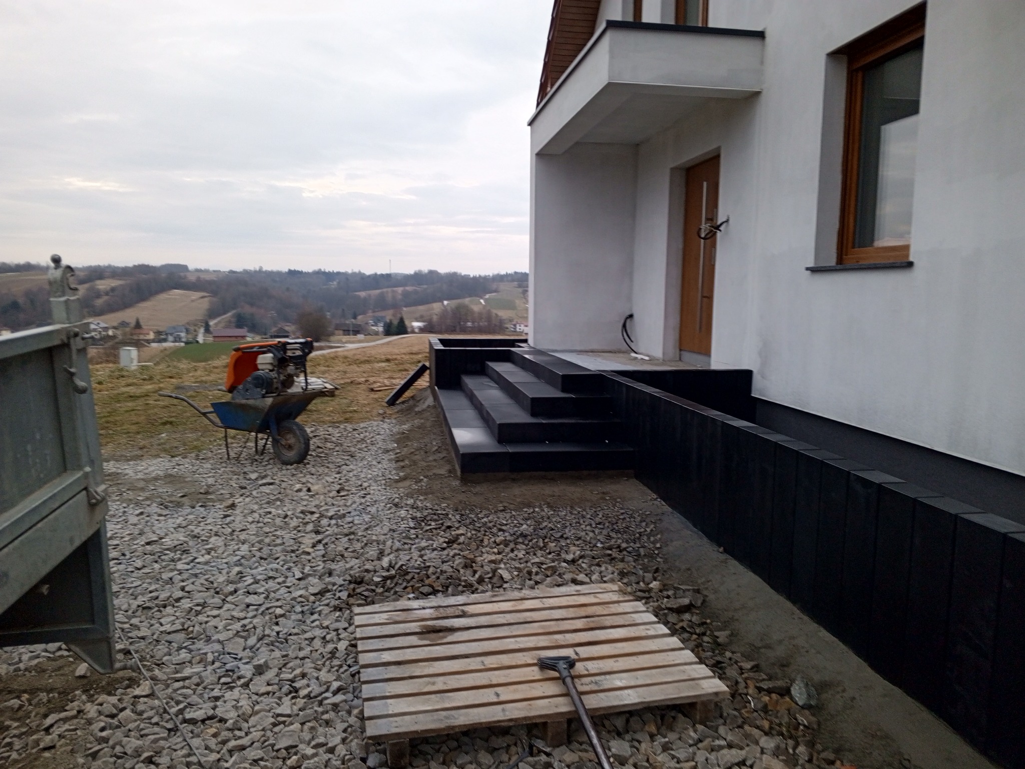 Nowoczesny dom z ciemnymi schodami wejściowymi i obłożoną płytami elewacją, widok na dolinę, taczka z zagęszczarką i paleta z narzędziem na żwirowym podłożu.