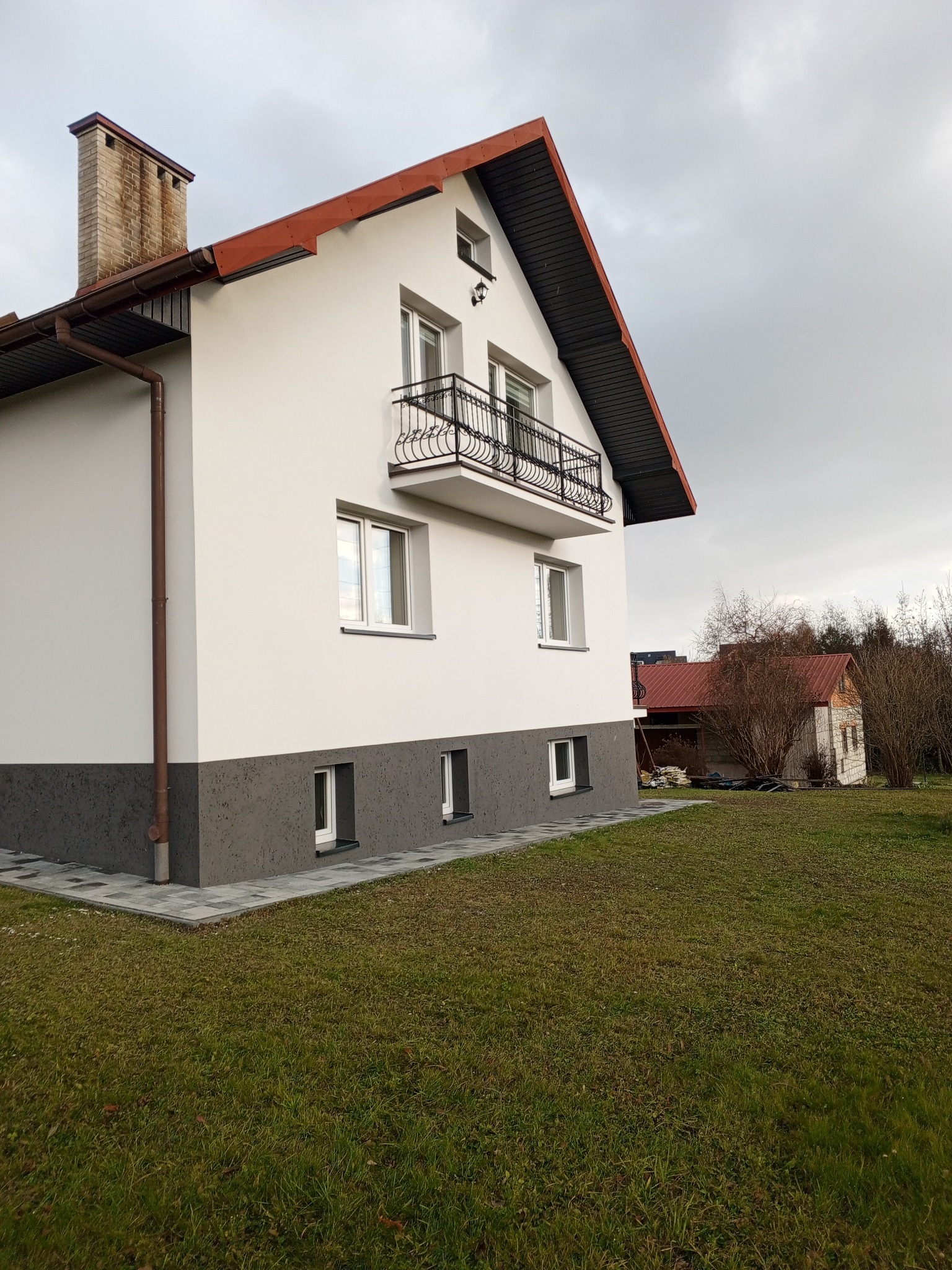 Świeżo otynkowany, dwukondygnacyjny dom z balkonem i nową elewacją w odcieniach bieli i szarości, z widocznym kominem z cegły i brązowymi rynnami na tle pochmurnego nieba.