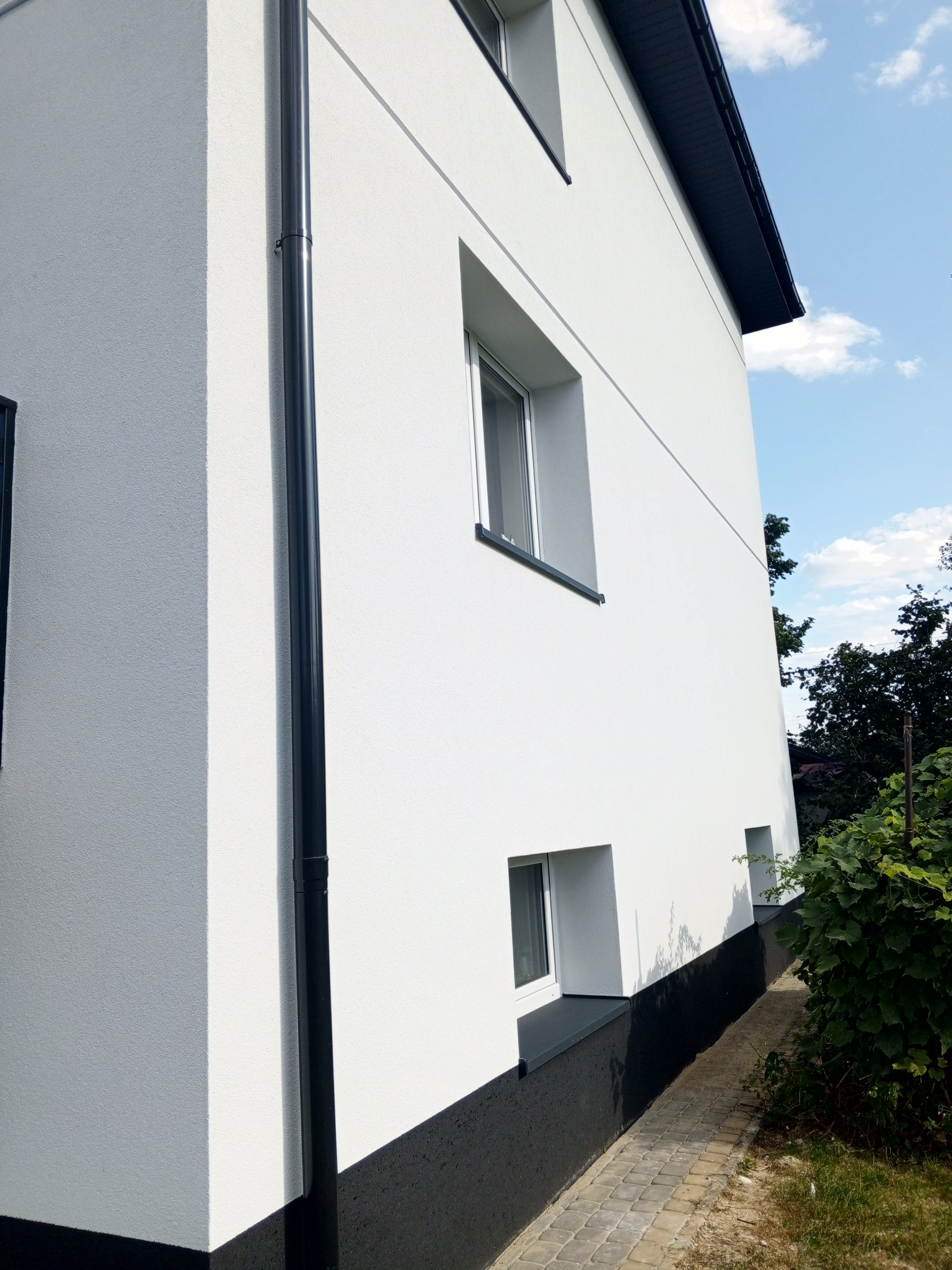 Boczny widok otynkowanego na biało domu z czarną rynną, cokołem i obramowaniami okien, widoczne okna o różnych rozmiarach i chodnik z kostki brukowej.