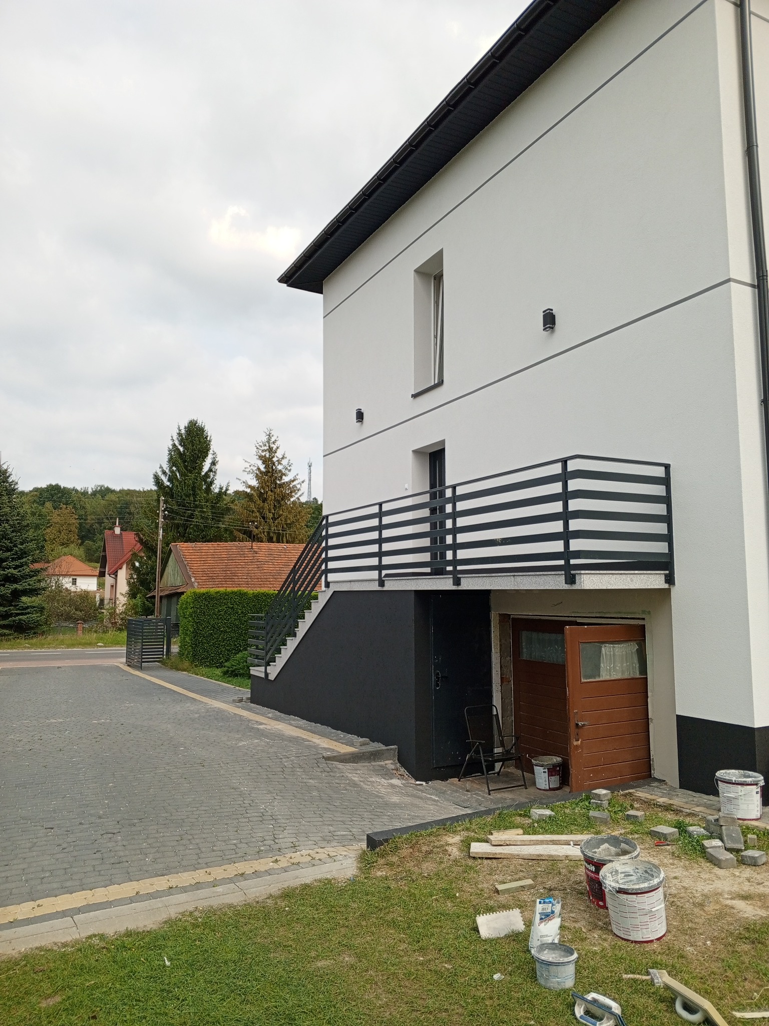 Dwupiętrowy dom z białymi ścianami i czarnymi akcentami, widok z zewnątrz na fragment elewacji, balkon z czarną balustradą, schody zewnętrzne, otwarte drzwi garażowe, puszki z farbą i narzędzia...