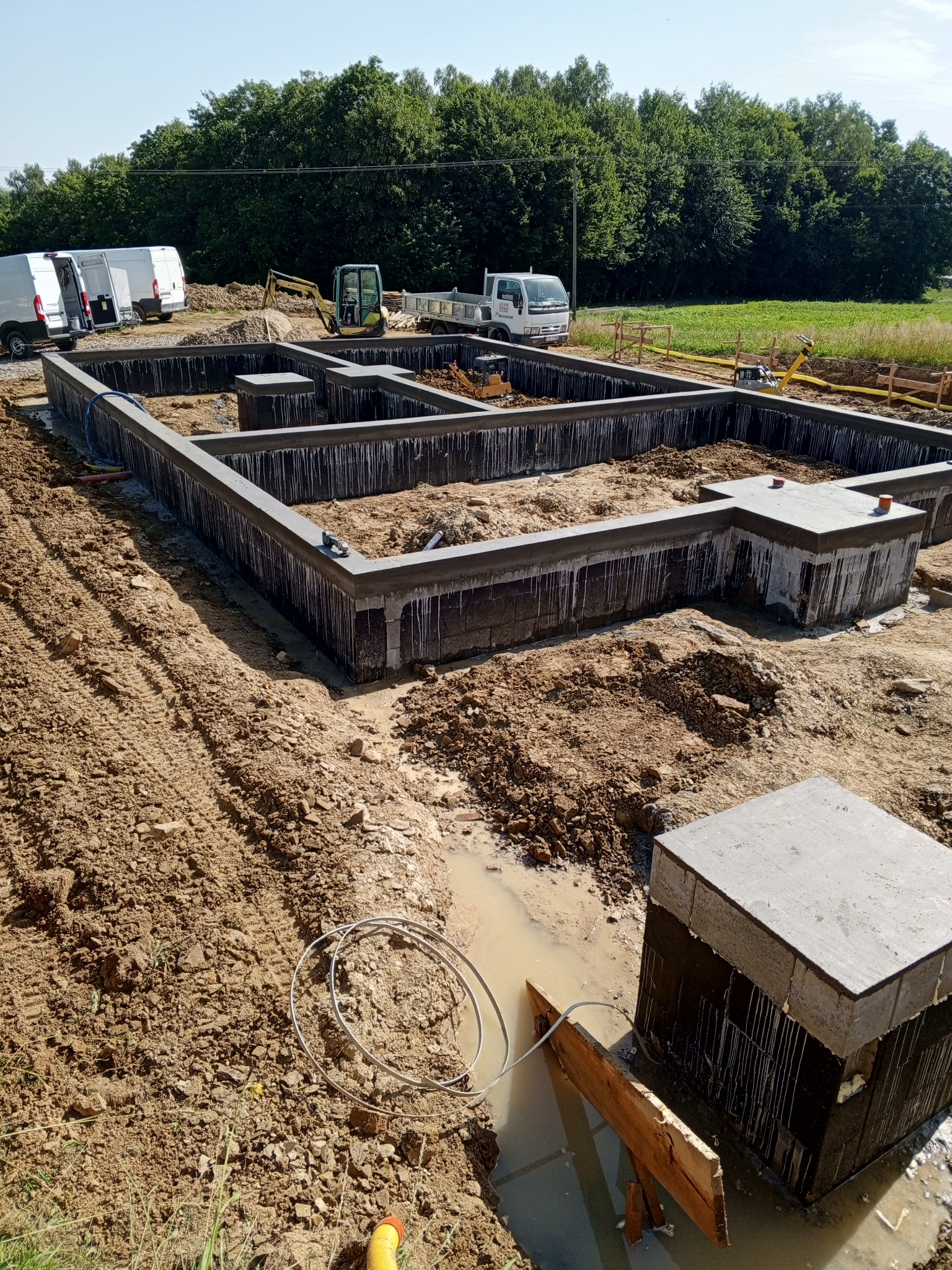 Wykop pod fundamenty domu jednorodzinnego z widocznymi zbrojeniami, deskowaniem i izolacją przeciwwilgociową, na działce budowlanej z zaparkowanymi pojazdami i drzewami w tle.