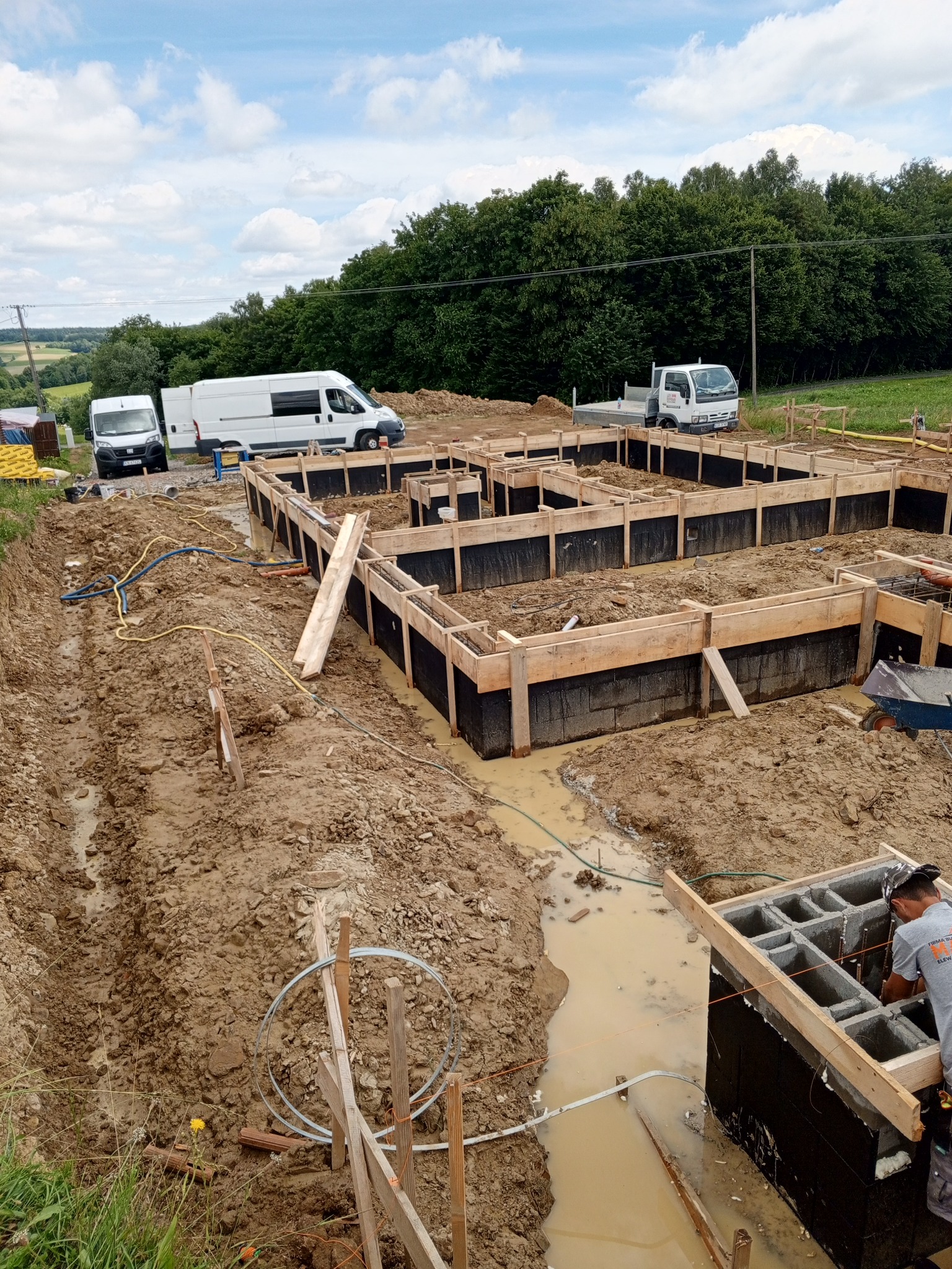 Budowa fundamentów domu: szalunki zalane wodą, widoczne zbrojenia, pracownik w szarej koszulce, dwa busy i ciężarówka w tle, teren budowy w błocie, pochmurne niebo.
