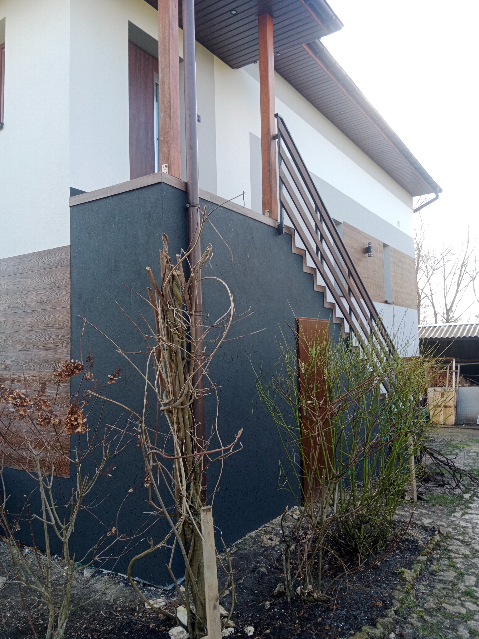 Dwukondygnacyjny dom z zewnętrznymi schodami z drewnianą balustradą, wykończony w tonacji bieli, szarości i drewna, z widocznym fragmentem elewacji o fakturze drewna i ciemnoszarą ścianą, otoczony...