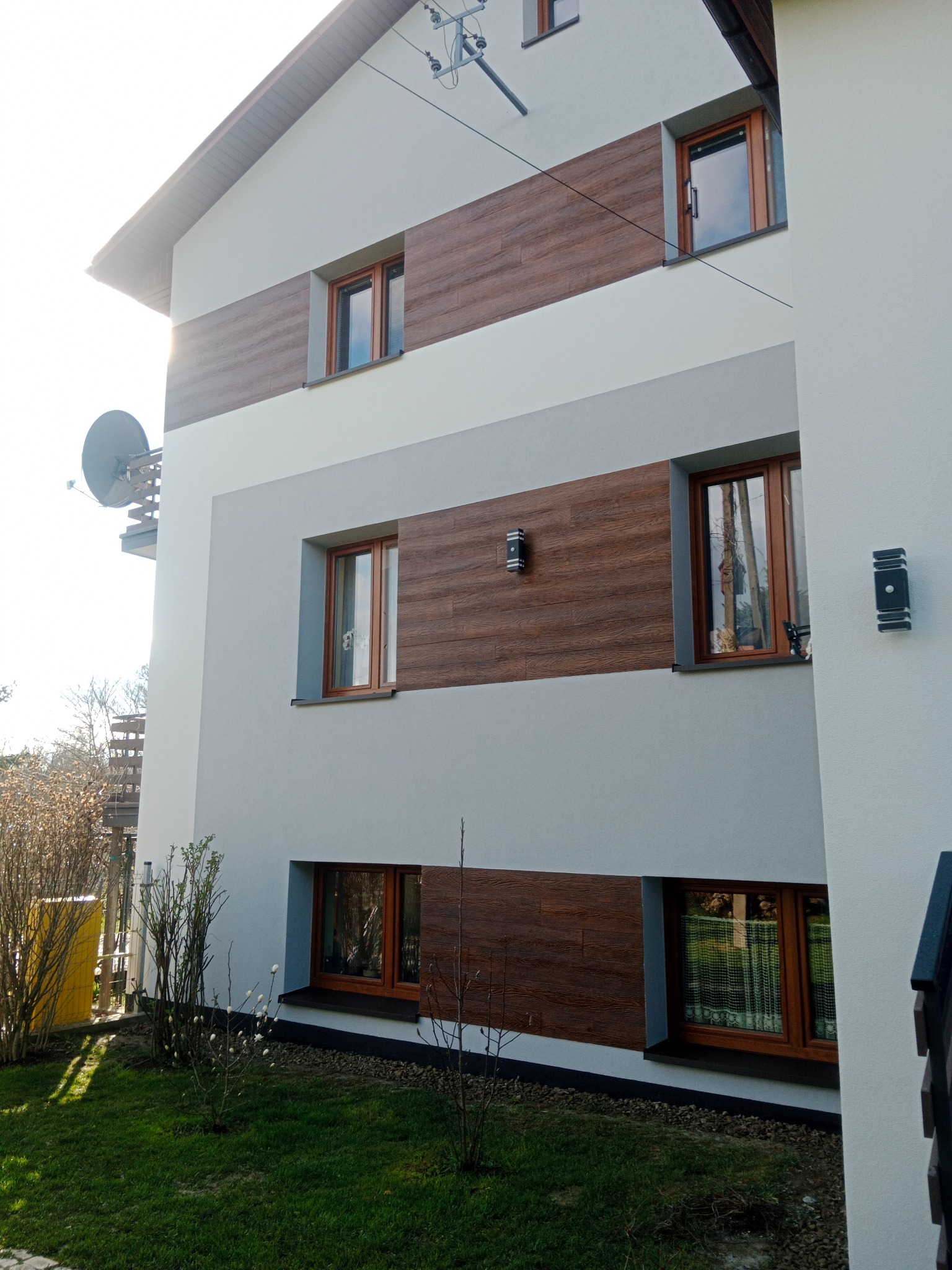 Nowoczesny dom z elewacją w odcieniach szarości i brązu, z oknami w drewnianych ramach, oświetleniem zewnętrznym i anteną satelitarną na balkonie.