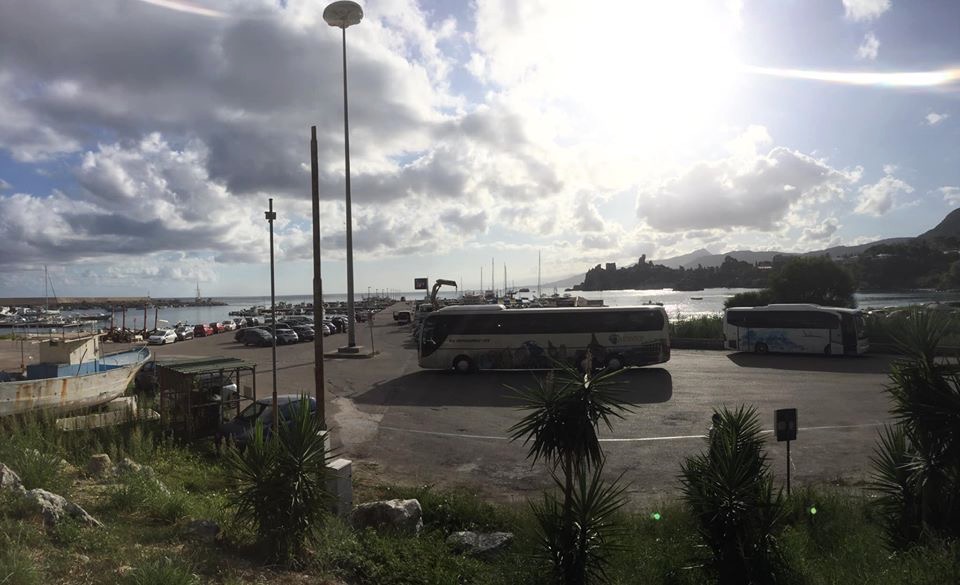 Dwa autokary na parkingu portowym z widokiem na morze i górzysty krajobraz w tle, pod pochmurnym niebem z przebłyskami słońca.