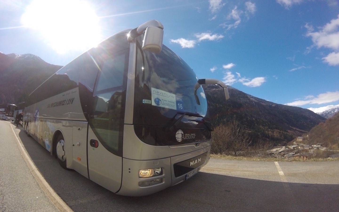 Nowoczesny autokar turystyczny MAN z logo Alextour zaparkowany na tle górskiego krajobrazu w słoneczny dzień.