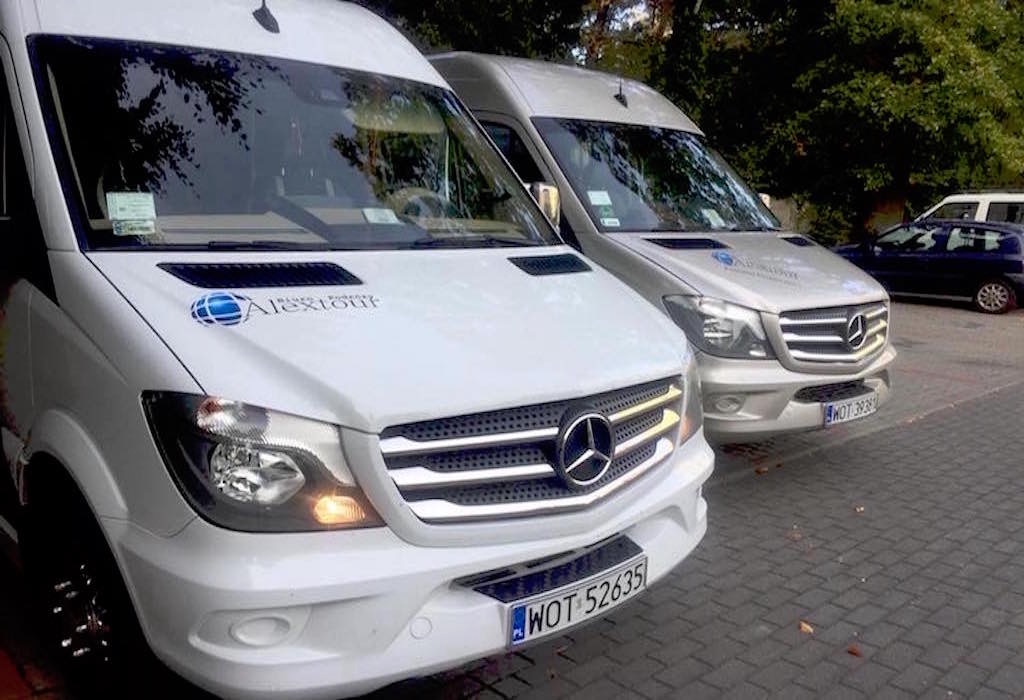 Dwa busy Mercedes-Benz Sprinter, biały i srebrny, z logo firmy przewozowej 'Alextour' na masce, zaparkowane na brukowanym parkingu.