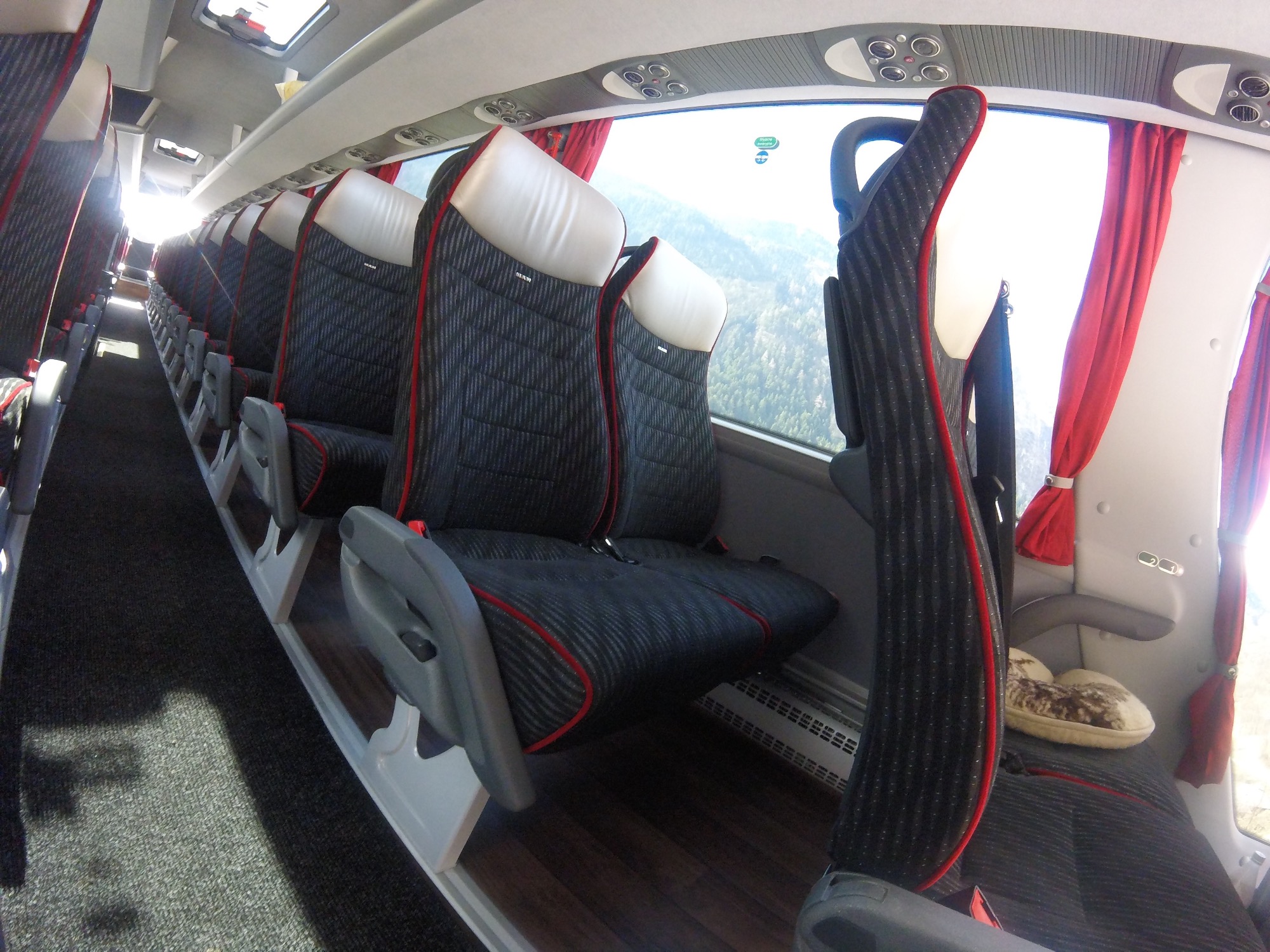 Wnętrze autobusu turystycznego z rzędem pustych, tapicerowanych foteli z czerwonymi akcentami, widok na krajobraz górski za oknem, kapcie na jednym z siedzeń.