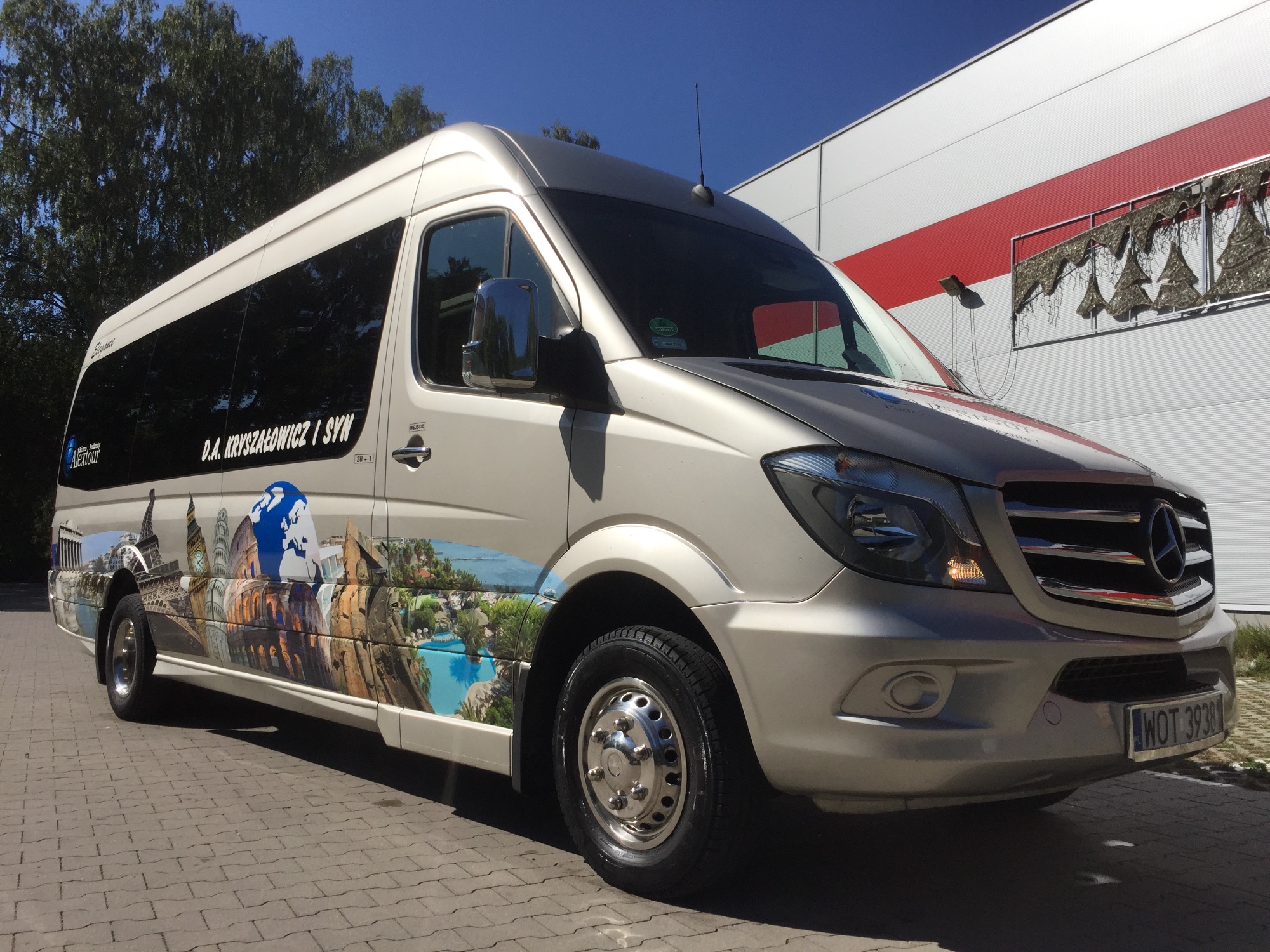 Szary bus marki Mercedes z grafiką przedstawiającą znane budowle świata, zaparkowany na brukowanej powierzchni w słoneczny dzień.