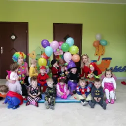 Grupa dzieci w przebraniach, siedzących na podłodze w sali z zielonymi ścianami, w tle dwie kobiety w perukach i strojach klauna oraz dekoracje z balonów.