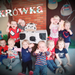 Żłobek Miniodkrywcy - Grupa uśmiechniętych dzieci w różnym wieku siedzi i macha rękami w sali ozdobionej motywami zwierzęcymi i napisem 'Krowka' wykonanym z kwiatowych liter, za nimi dekoracja imitująca krowę z kartonu.