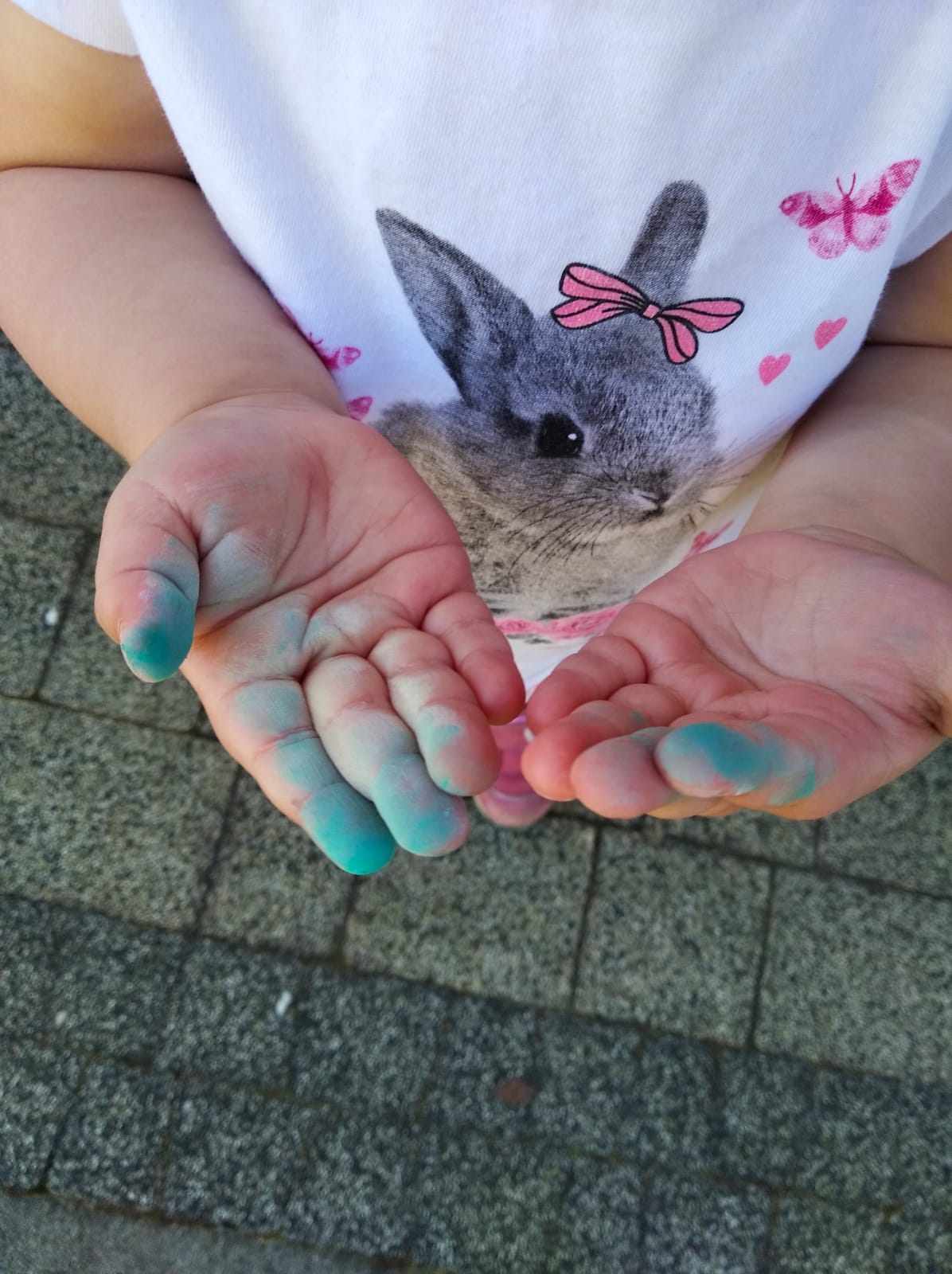 Dziecięce dłonie z plamami jasnoniebieskiej farby, wyciągnięte do przodu na tle chodnika i koszulki z nadrukiem królika z różową kokardką.