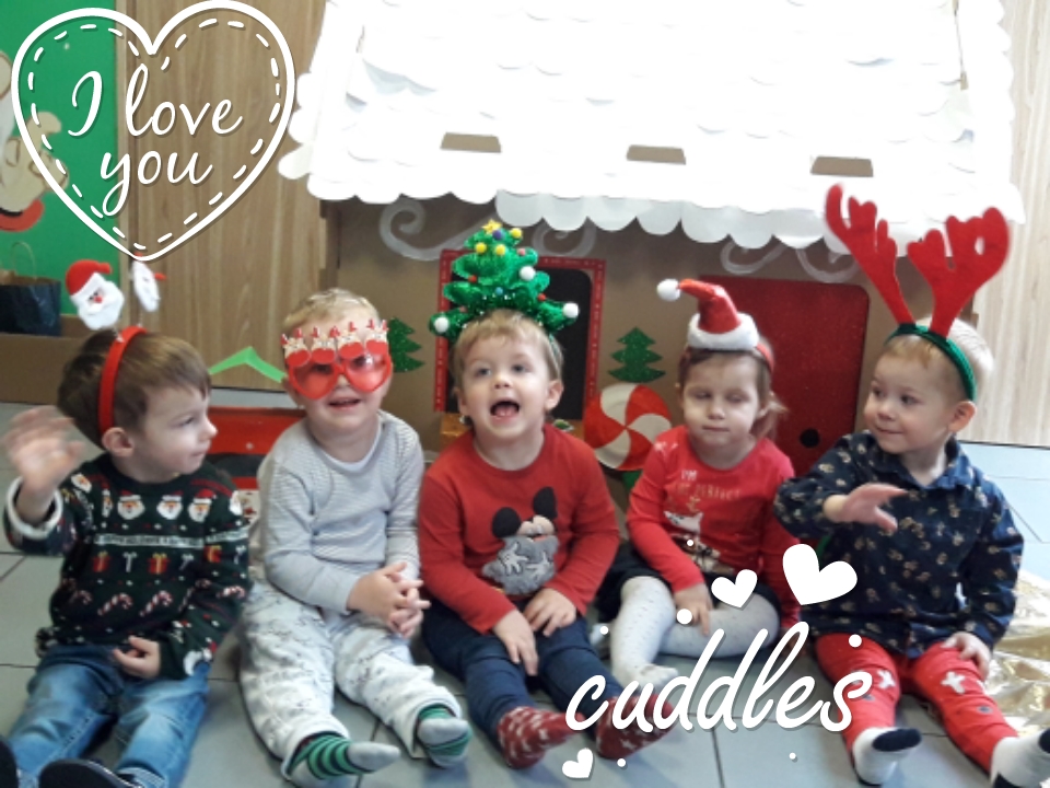 Pięcioro uśmiechniętych dzieci siedzi na podłodze przed kartonową dekoracją przypominającą domek z piernika, ubrani w świąteczne stroje i opaski z motywami świątecznymi, z dodanymi cyfrowo...