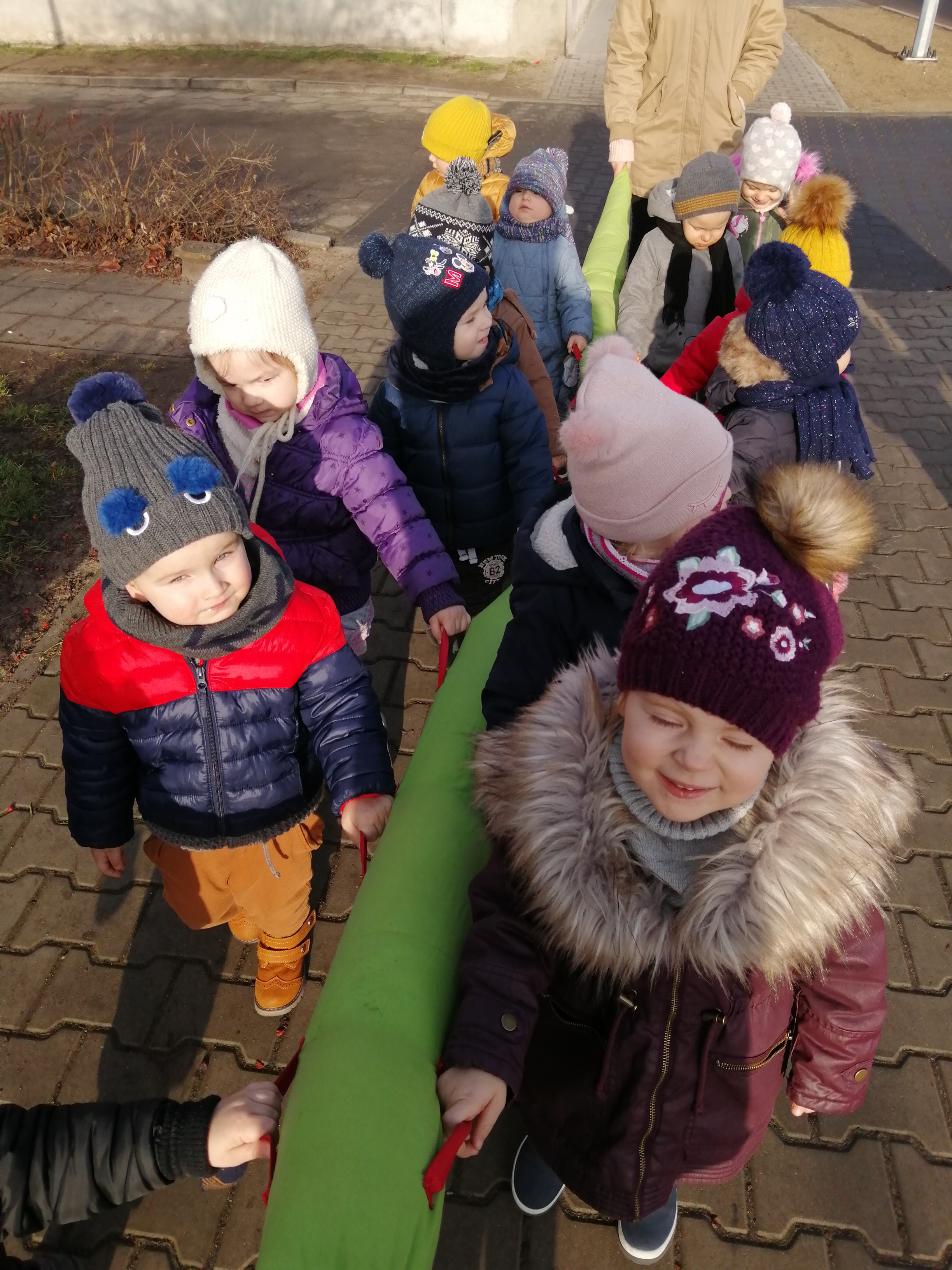 Grupa dzieci w zimowych ubraniach trzymająca się zielonej, podłużnej poduszki z czerwonymi uchwytami na zewnątrz, być może podczas spaceru lub zabawy.