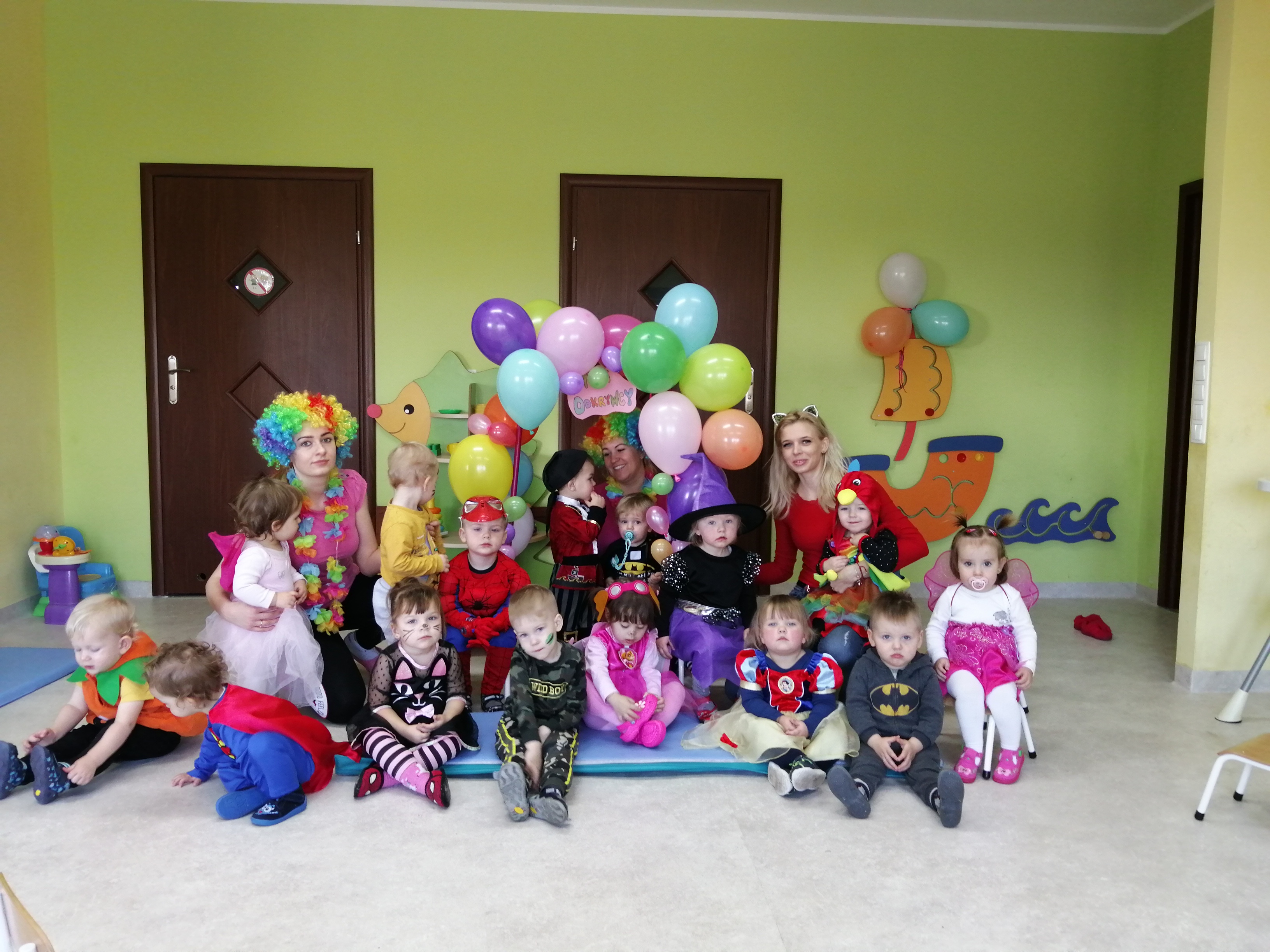 Grupa dzieci w przebraniach, siedzących na podłodze w sali z zielonymi ścianami, w tle dwie kobiety w perukach i strojach klauna oraz dekoracje z balonów.