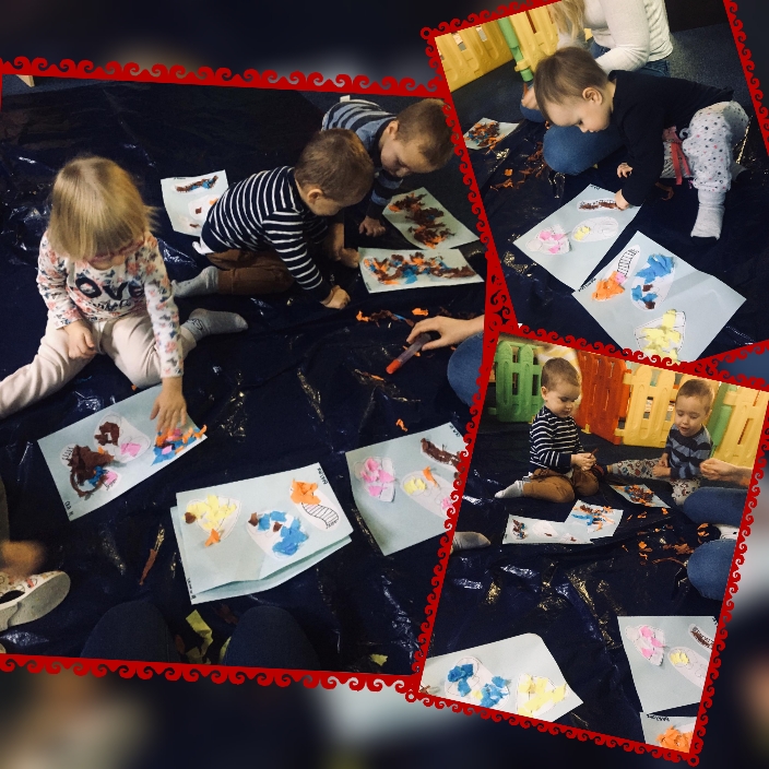 Grupa dzieci siedzi na czarnej folii, tworząc kolaże z kolorowych papierowych elementów, używając kleju w sztyfcie, pod nadzorem osoby dorosłej.