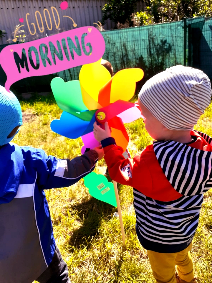 Dwoje dzieci bawi się kolorowym wiatraczkiem na trawie w słoneczny dzień, z efektem graficznym 'Good Morning' w rogu.