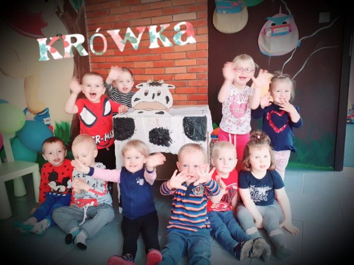 Grupa uśmiechniętych dzieci w różnym wieku siedzi i macha rękami w sali ozdobionej motywami zwierzęcymi i napisem 'Krowka' wykonanym z kwiatowych liter, za nimi dekoracja imitująca krowę z kartonu.