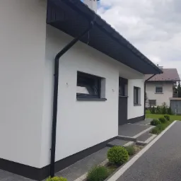 Boczny widok nowo otynkowanego domu z czarnymi detalami: rynny, obramowania okien, cokół. Minimalistyczny styl, jasna elewacja, szara kostka brukowa i ozdobne krzewy.