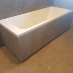 Slawko-bud - Nowoczesna, prostokątna wanna obudowana szarą płytką imitującą beton, z widocznymi przyłączami na ścianie i brązową podłogą z płytek. Minimalistyczny design łazienki.