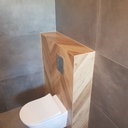 Slawko-bud - Podwieszana toaleta z zabudową w jodełkę, szara płytka ścienna i podłogowa. Nowoczesna aranżacja łazienki z akcentem drewna i minimalistycznym przyciskiem spłukującym.