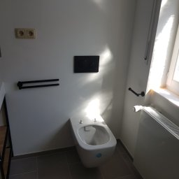 Slawko-bud - Nowoczesna toaleta podwieszana w jasnej łazience z szarymi płytkami podłogowymi. Widoczne akcesoria: uchwyt na ręcznik, przycisk spłuczki, grzejnik.