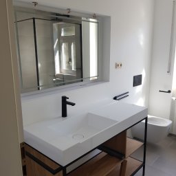 Slawko-bud - Nowoczesna łazienka z białą umywalką nablatową na drewnianym stelażu z czarnymi elementami, lustro w ramie i szara podłoga. Widoczna toaleta podwieszana.