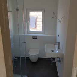 Slawko-bud - Nowoczesna łazienka w trakcie wykończenia: biała armatura (umywalka, toaleta), szklana kabina prysznicowa, ciemna podłoga. Widoczne niedokończone elementy instalacji.