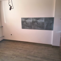 Slawko-bud - Jasny pokój po remoncie: drewnopodobne panele na podłodze, szare płytki imitujące marmur na ścianie, różowa ściana, widoczna instalacja oświetleniowa.