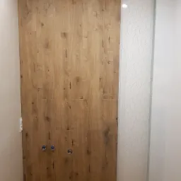 Wnętrze łazienki w trakcie prac wykończeniowych, widoczna ściana wyłożona płytami imitującymi drewno, obok fragment ściany z wzorzystymi białymi płytkami, widoczne wystające rury i instalacja...