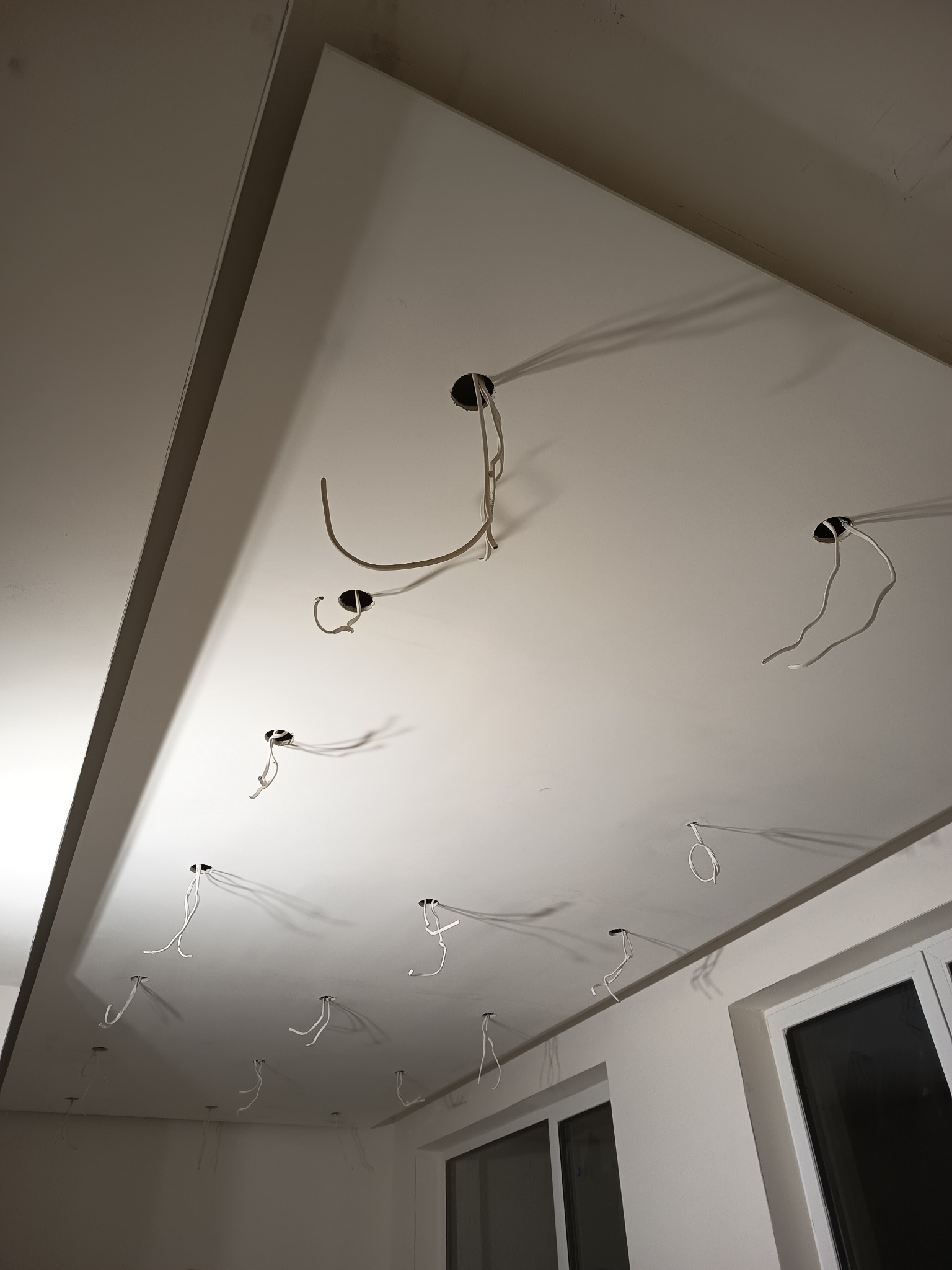 Biały sufit podwieszany z wystającymi kablami elektrycznymi z otworów, przygotowany do montażu oświetlenia. Widoczne okno i fragment ściany.