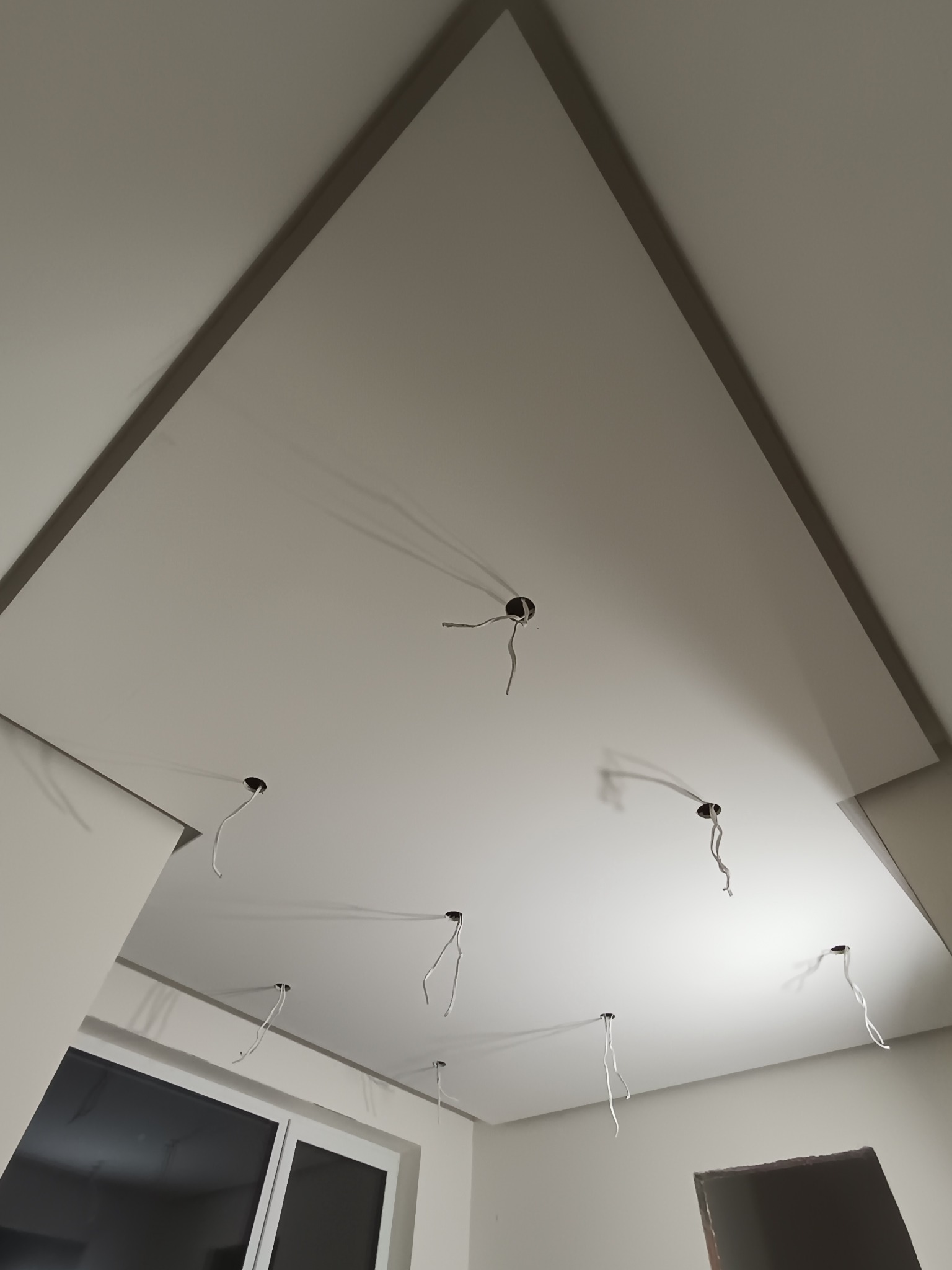 Podwieszany sufit z płyt karton-gips z widocznymi otworami na oświetlenie i wystającymi przewodami elektrycznymi. Kątowe ujęcie z dołu.