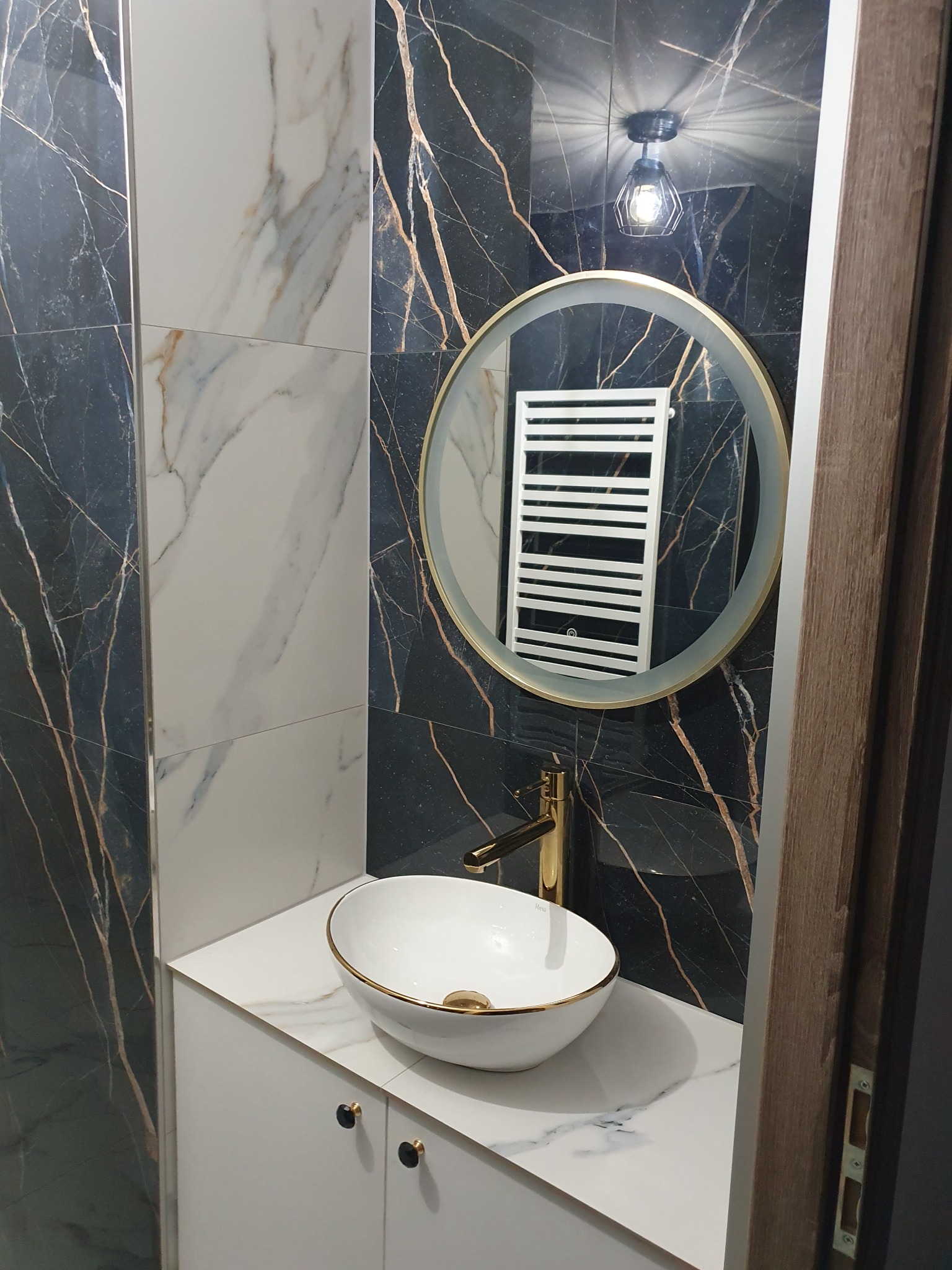 Elegancka łazienka z umywalką wpuszczaną w blat, złotą armaturą i okrągłym lustrem z podświetleniem. Ściany wyłożone płytkami imitującymi marmur w odcieniach bieli i czerni.