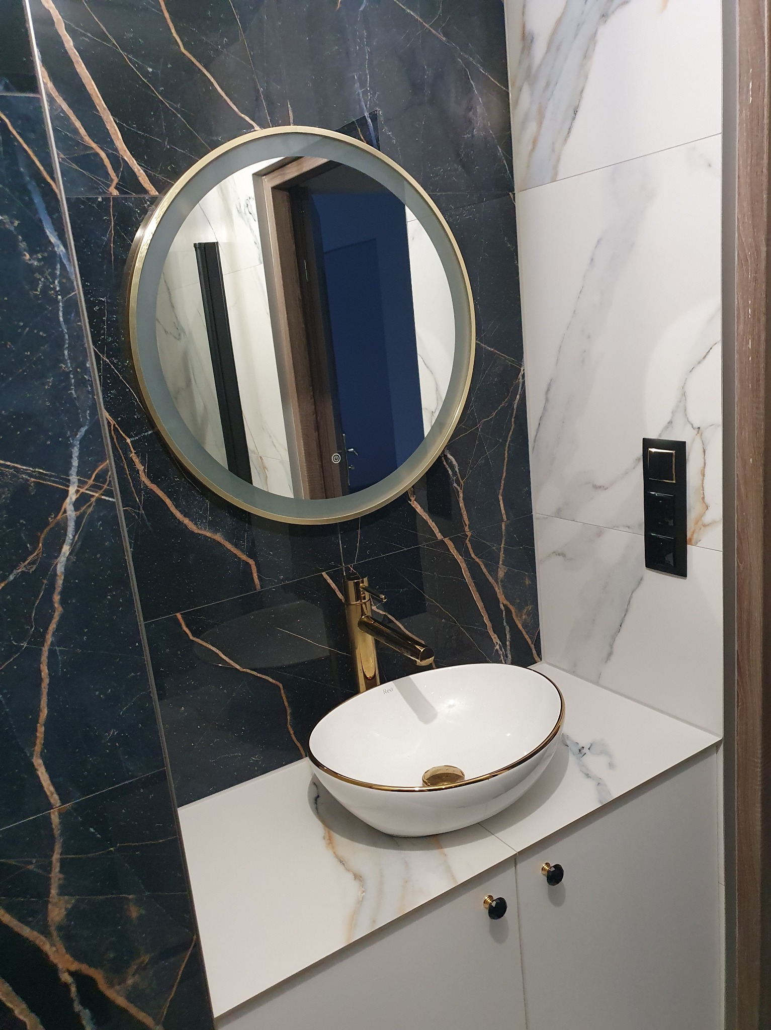 Elegancka łazienka z umywalką nablatową, złotą armaturą i okrągłym lustrem z podświetleniem. Ściany wyłożone płytkami imitującymi marmur w odcieniach bieli i granatu.