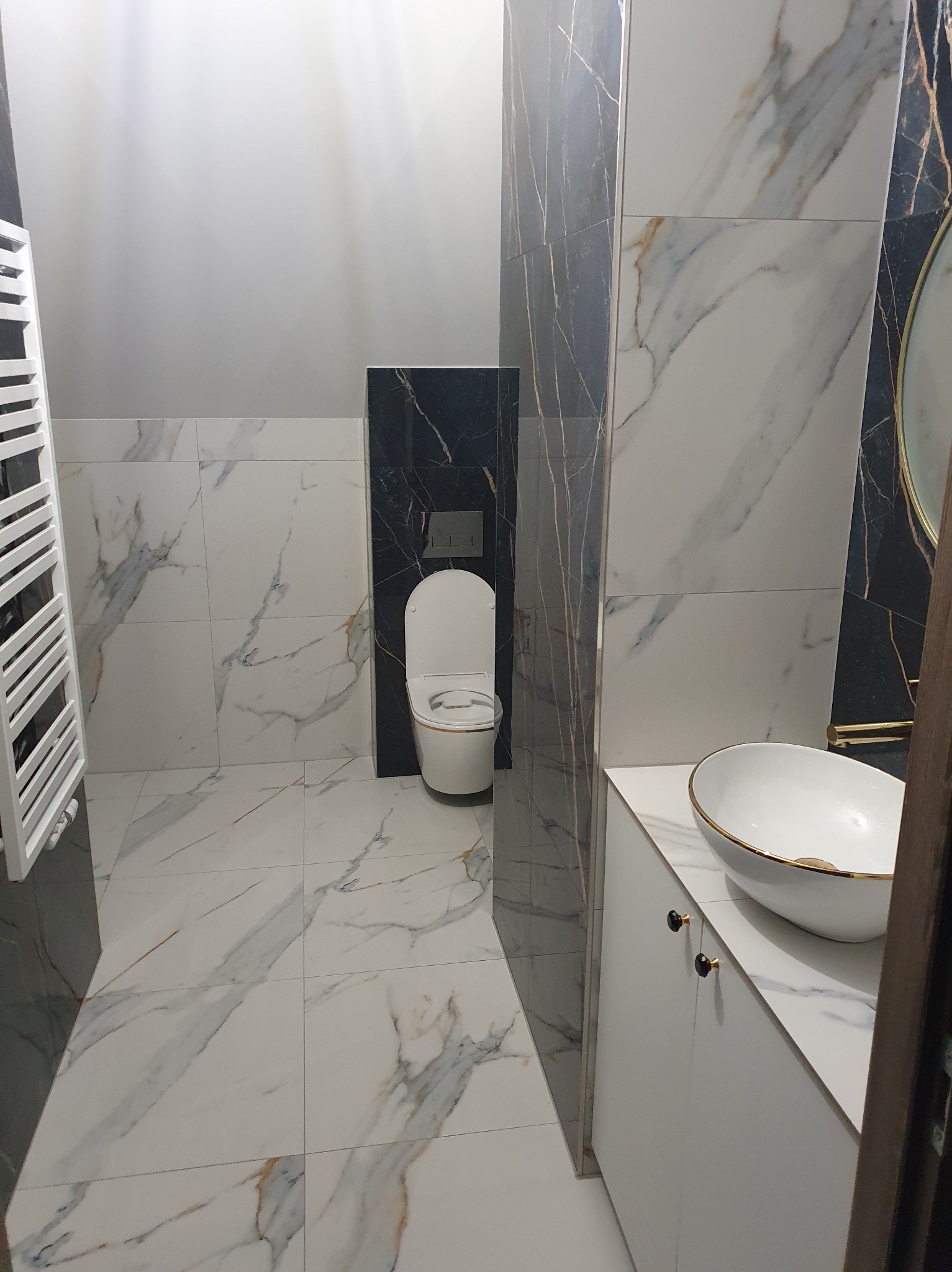 Wykończona łazienka w bieli i czerni z marmurkowym wzorem płytek. Umywalka nablatowa z złotym rantem, grzejnik drabinkowy i toaleta w tle.