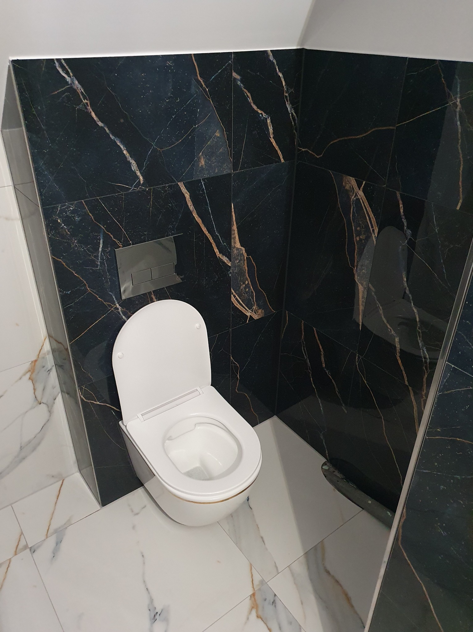Wykończona łazienka z czarnymi płytkami imitującymi marmur ze złotymi żyłami na ścianach i białymi płytkami na podłodze. Widoczna toaleta z otwartą klapą.