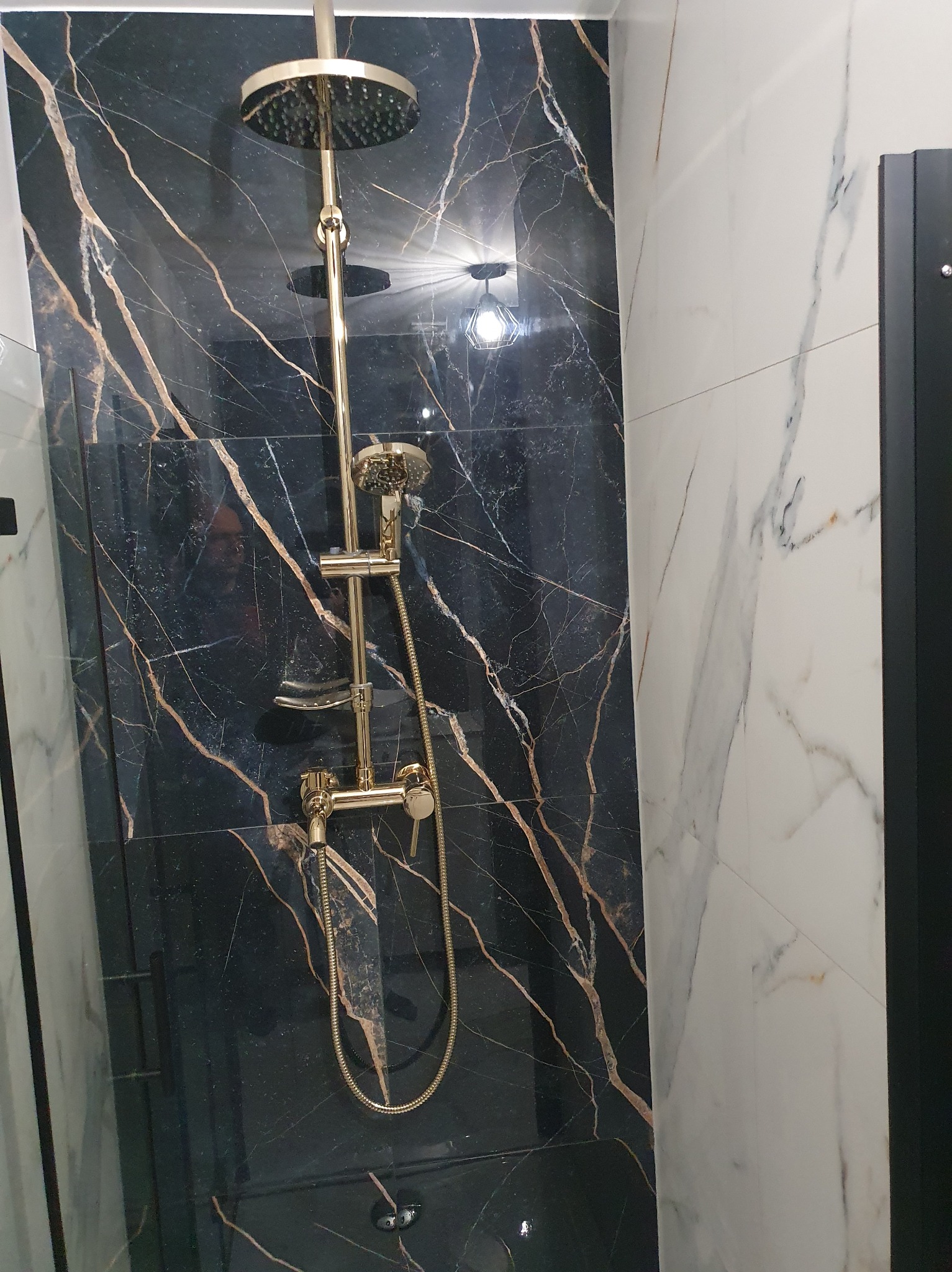 Luksusowa kabina prysznicowa z czarnymi płytkami imitującymi marmur z złotymi żyłami i złotą armaturą. Widoczne białe płytki imitujące marmur po bokach.