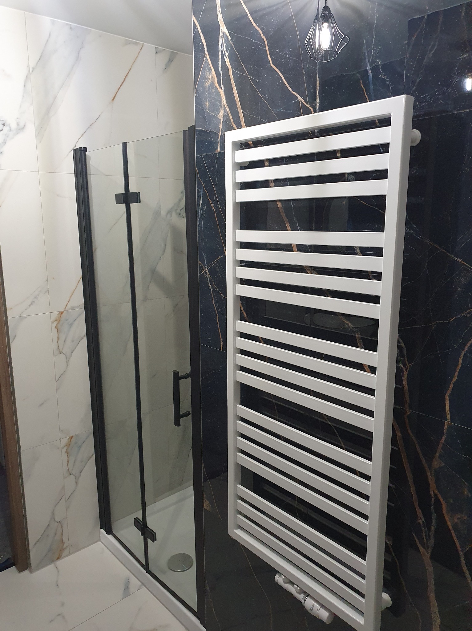 Nowoczesna łazienka z kabiną prysznicową typu walk-in z czarnymi profilami i białym grzejnikiem drabinkowym na tle czarnych płytek imitujących marmur.