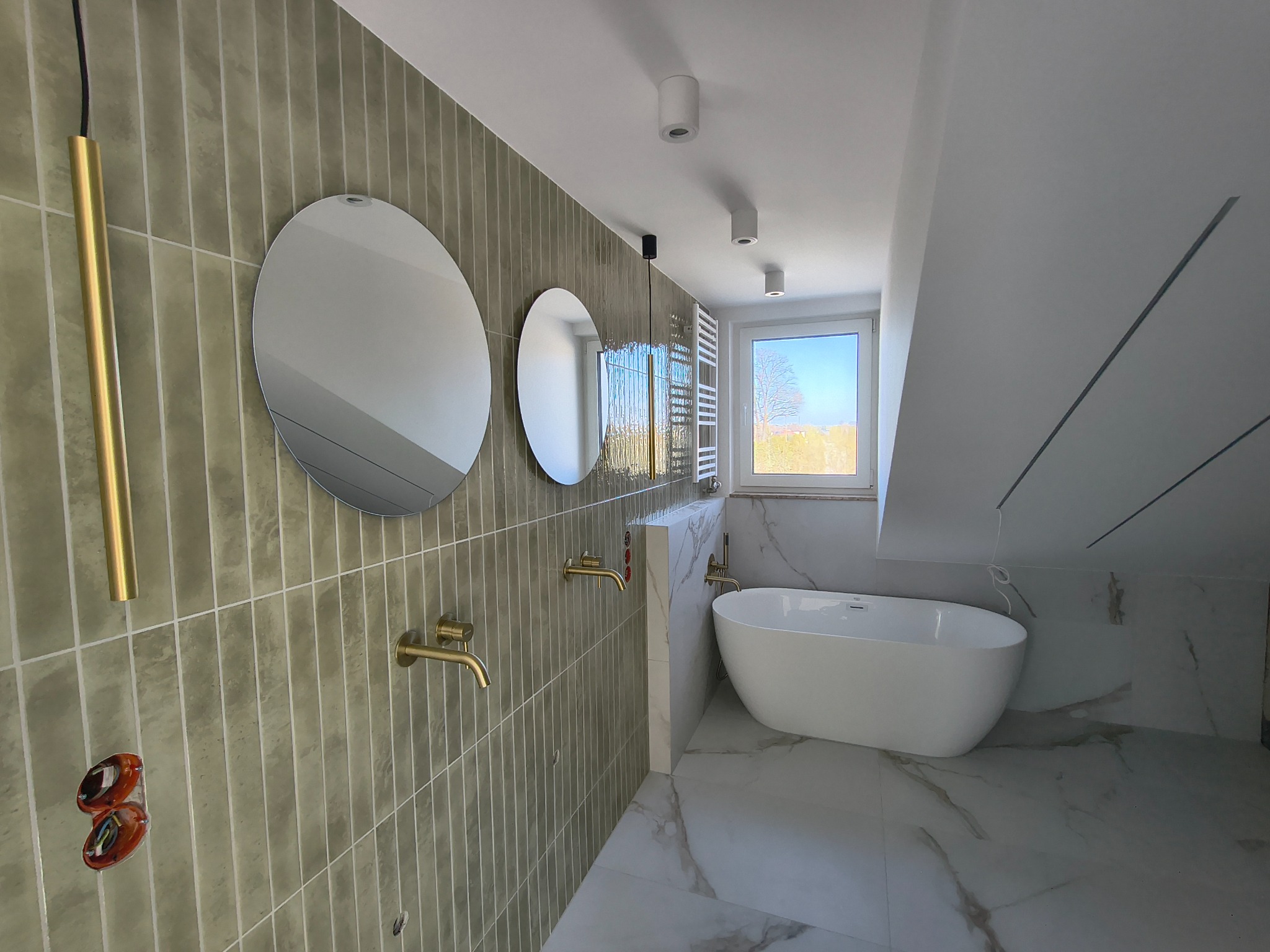 Nowoczesna łazienka na poddaszu z owalną wanną, okrągłymi lustrami i złotymi akcentami, wykończona płytkami w odcieniach szarości i bieli.