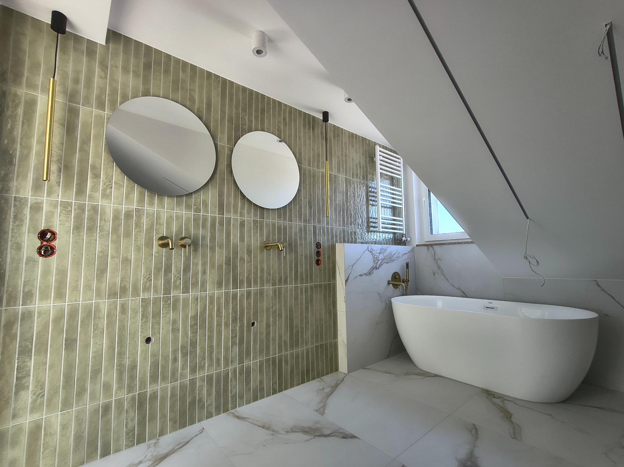 Nowoczesna łazienka na poddaszu z owalną wanną, marmurową podłogą i ścianami z podłużnych, oliwkowych płytek. Dwa okrągłe lustra i złote akcenty armatury.