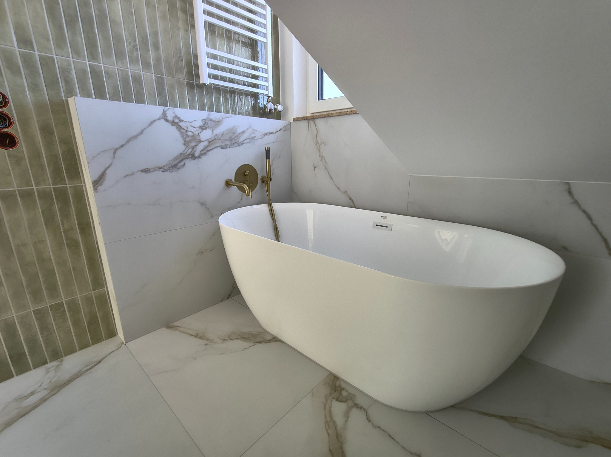 Nowoczesna łazienka na poddaszu z wolnostojącą białą wanną, złotą armaturą i marmurowymi płytkami. Uwagę przykuwają pionowe, oliwkowe płytki na ścianie.