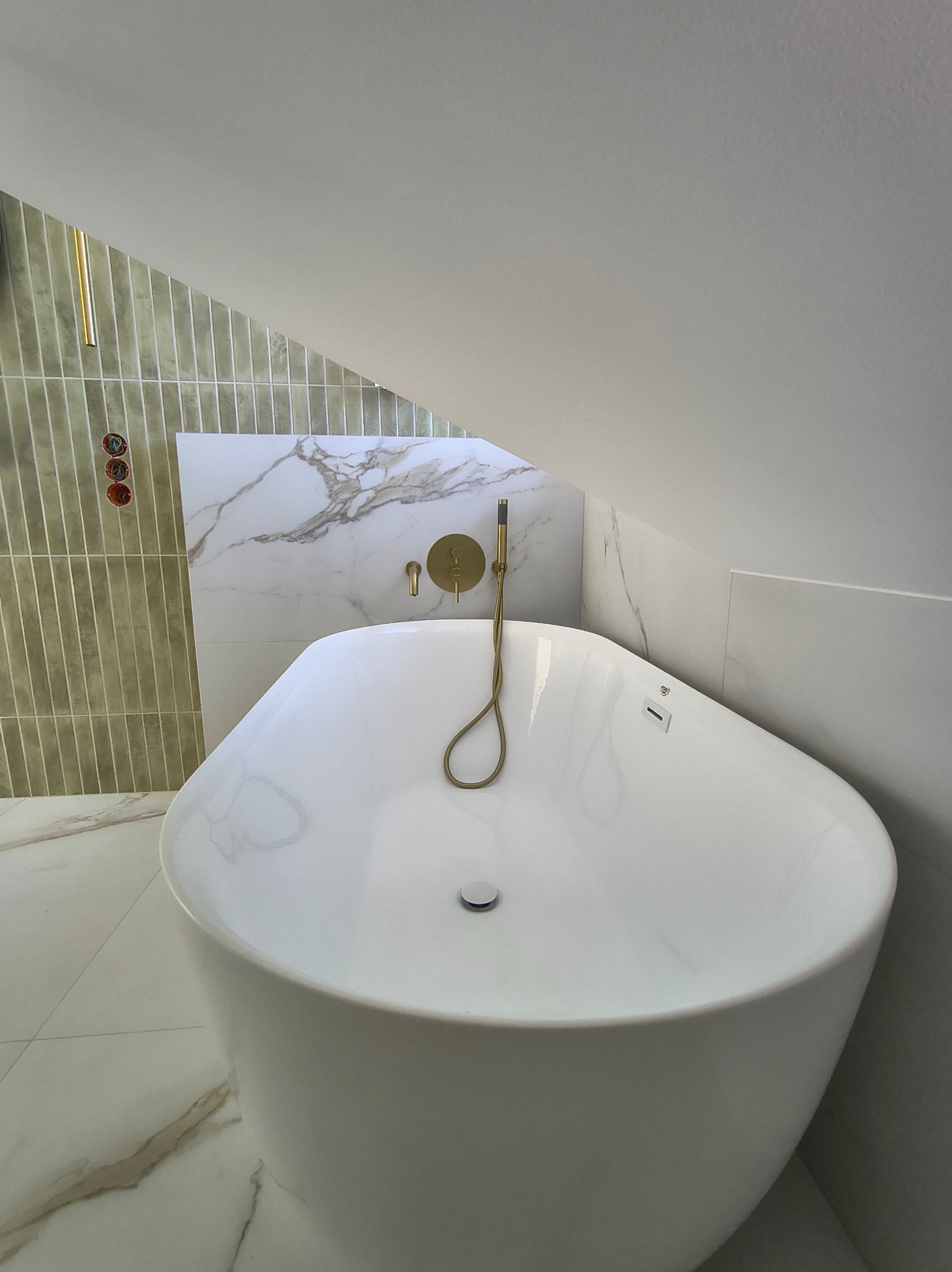 Nowoczesna, wolnostojąca wanna w łazience na poddaszu, z baterią w kolorze złotym. Ściany wyłożone płytkami imitującymi marmur i wąskimi, pionowymi płytkami.