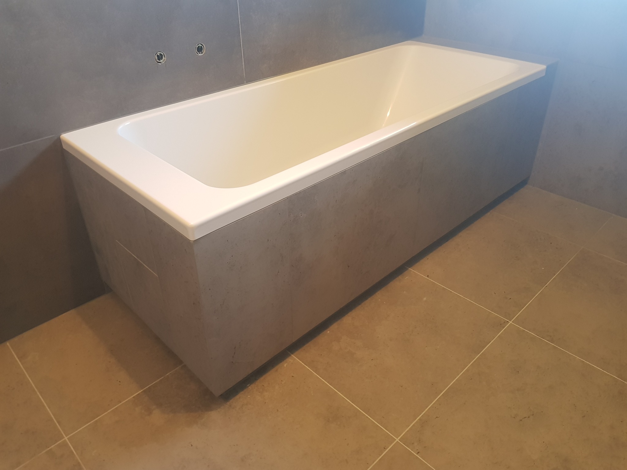 Nowoczesna, prostokątna wanna obudowana szarą płytką imitującą beton, z widocznymi przyłączami na ścianie i brązową podłogą z płytek. Minimalistyczny design łazienki.