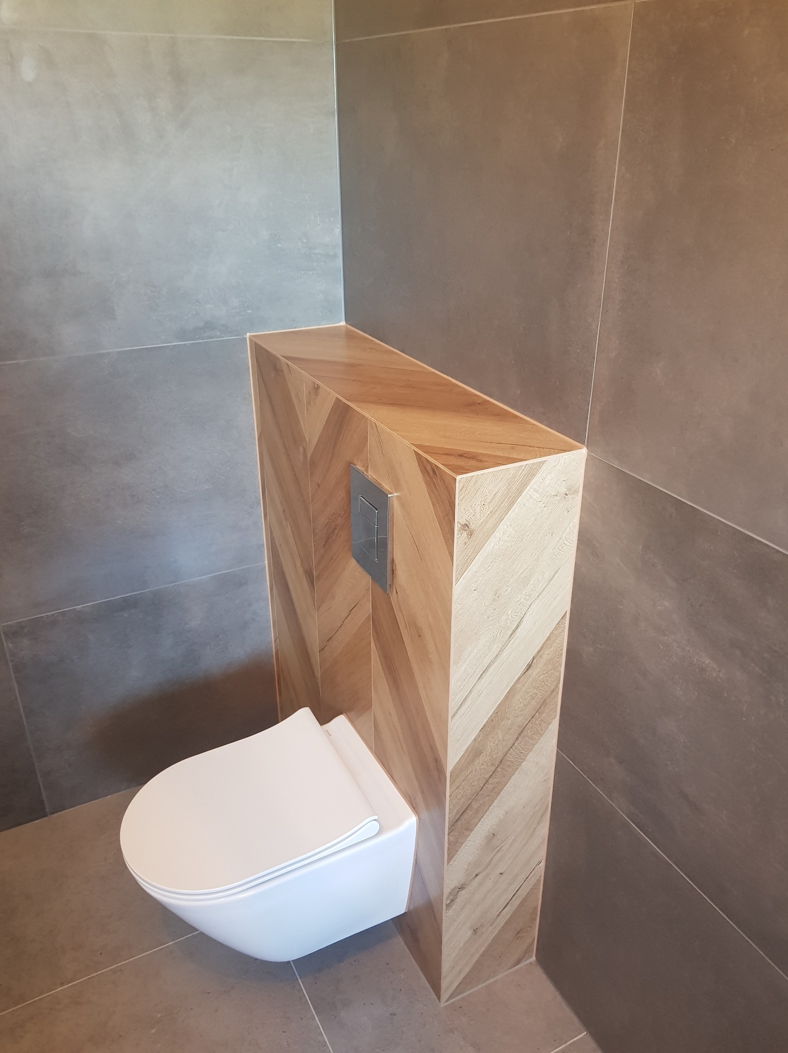 Podwieszana toaleta z zabudową w jodełkę, szara płytka ścienna i podłogowa. Nowoczesna aranżacja łazienki z akcentem drewna i minimalistycznym przyciskiem spłukującym.