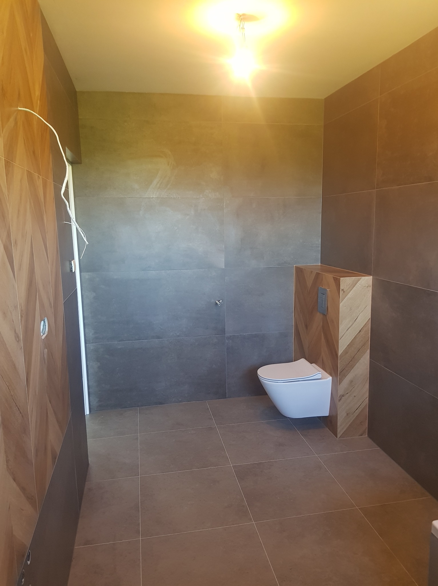 Nowoczesna łazienka z szarymi płytkami i drewnianą obudową toalety podtynkowej. Uwagę zwraca wzór jodełki na obudowie i minimalistyczna armatura.