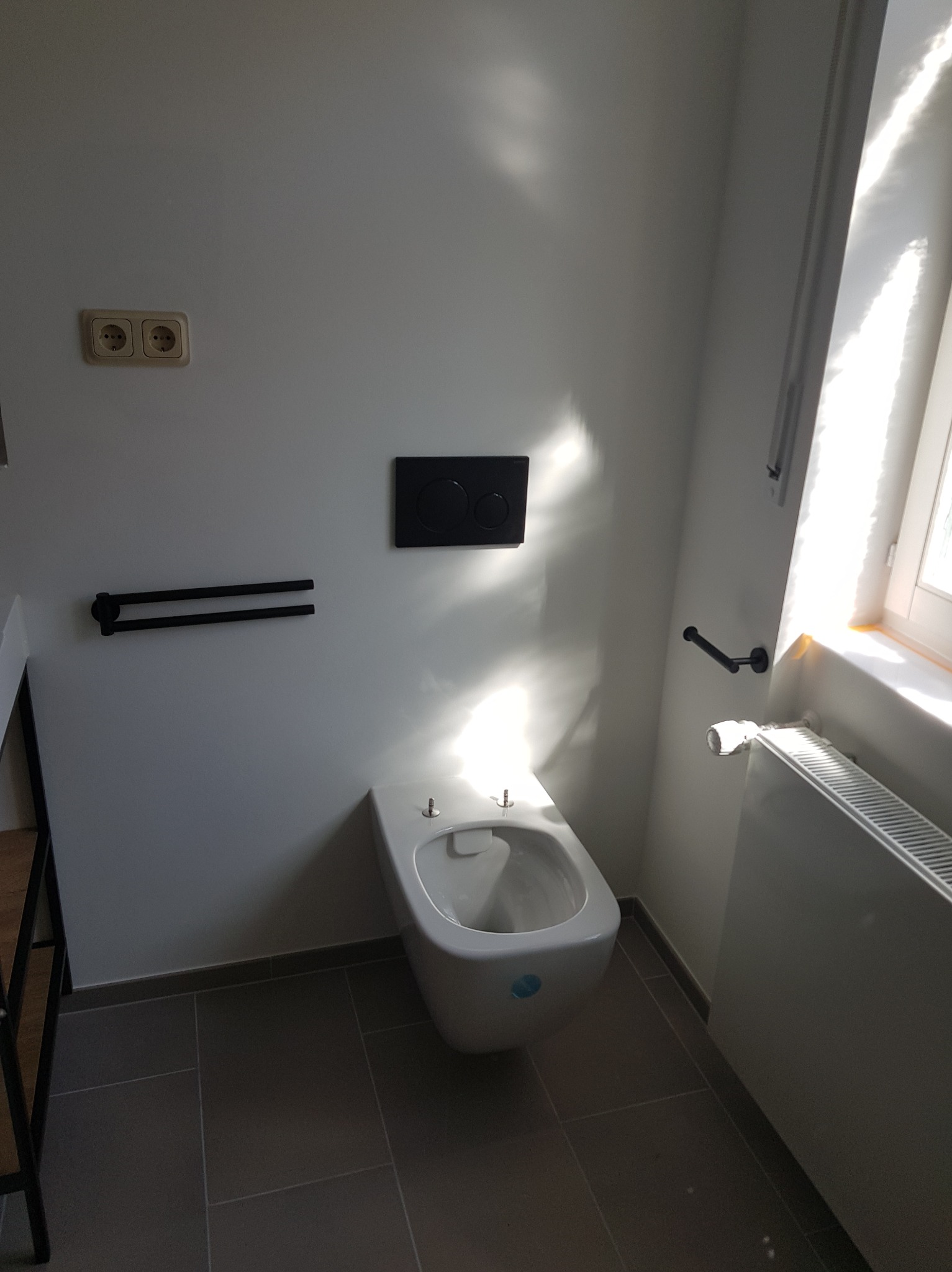 Nowoczesna toaleta podwieszana w jasnej łazience z szarymi płytkami podłogowymi. Widoczne akcesoria: uchwyt na ręcznik, przycisk spłuczki, grzejnik.