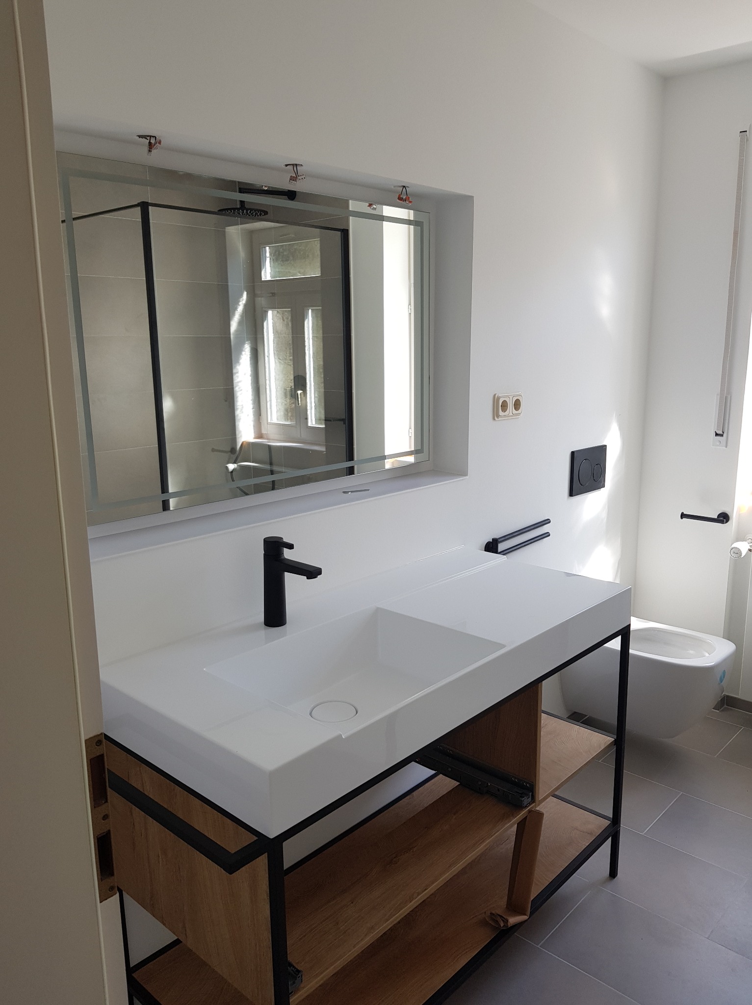 Nowoczesna łazienka z białą umywalką nablatową na drewnianym stelażu z czarnymi elementami, lustro w ramie i szara podłoga. Widoczna toaleta podwieszana.